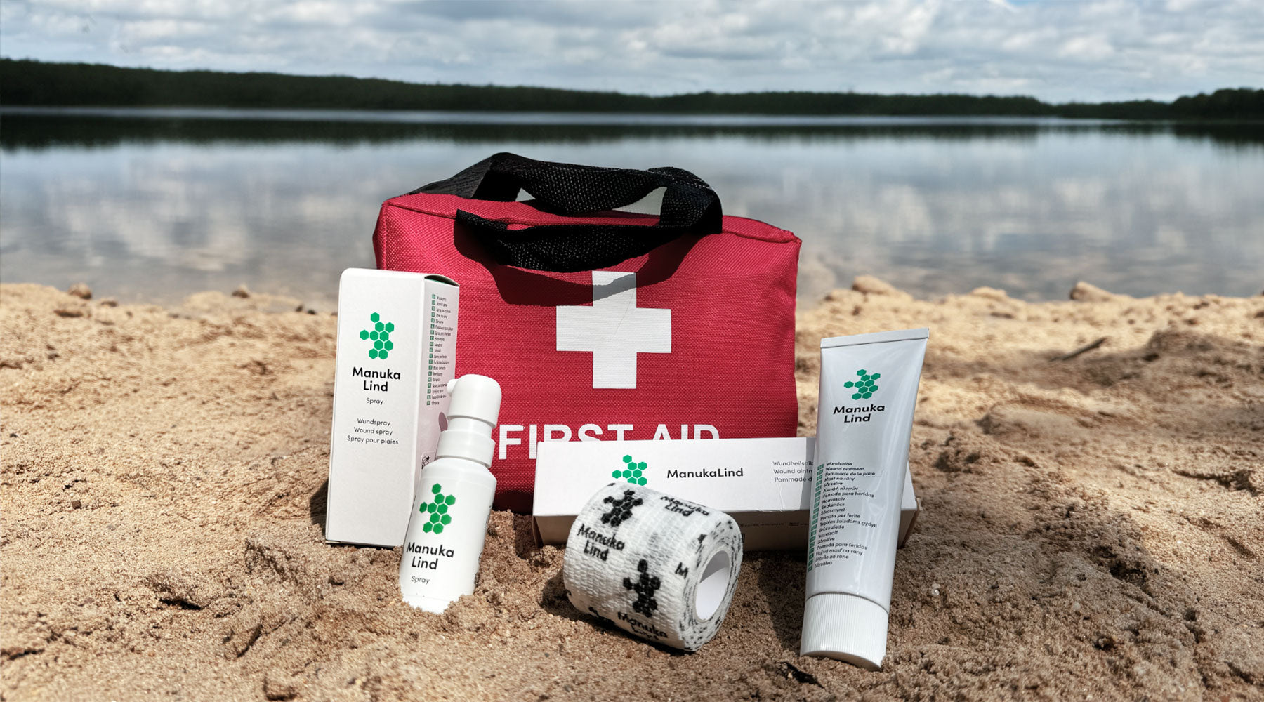 Ein Erste-Hilfe-Set mit ManukaLind-Produkten wird an einem Sandstrand neben einem ruhigen See präsentiert. Das Set umfasst ManukaLind Wundheilsalbe, Wundspray und Fixierandage