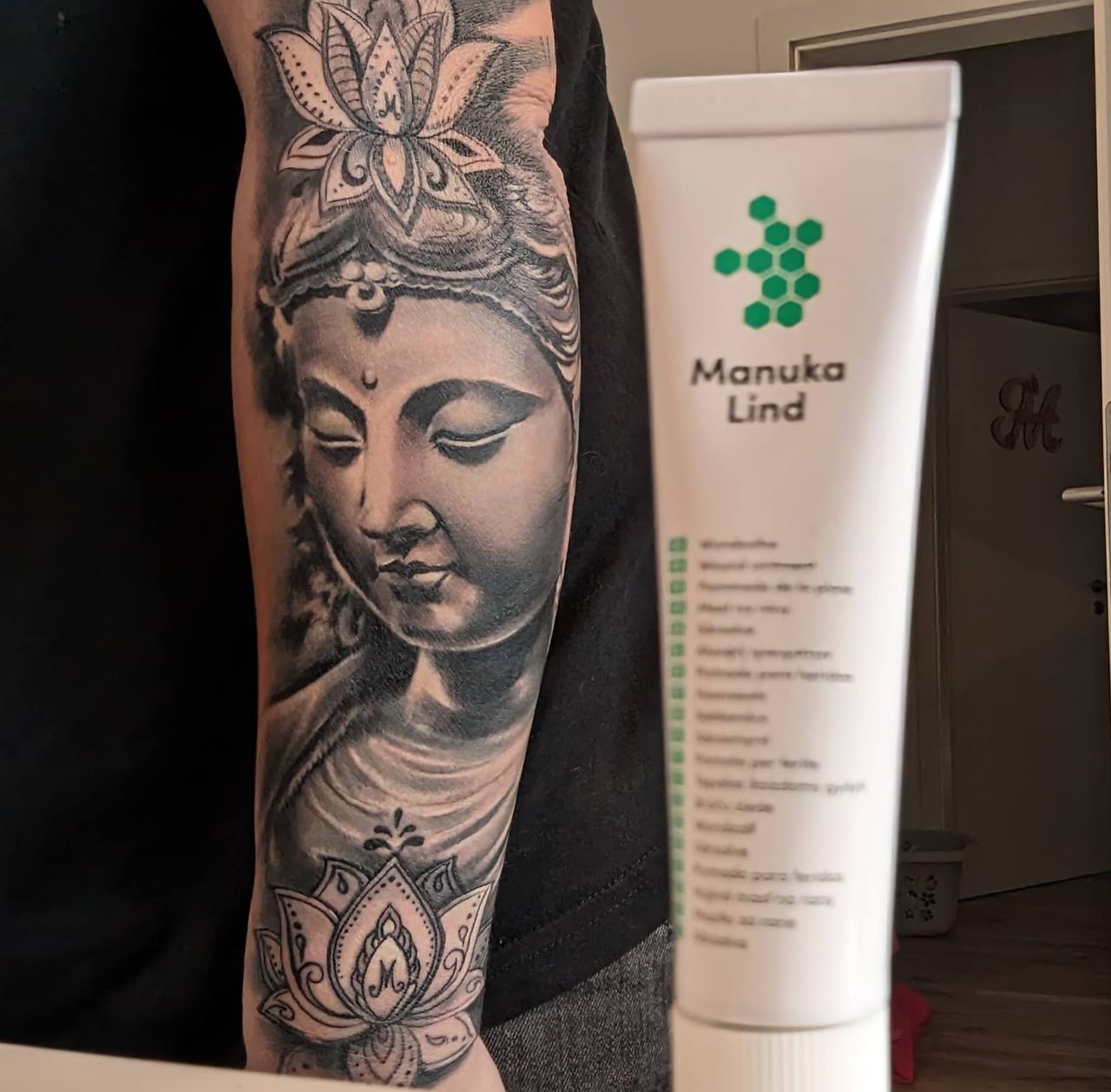 Ein tattoowierter Arm mit einer Statue neben einer Tube ManukaLind Salbe 